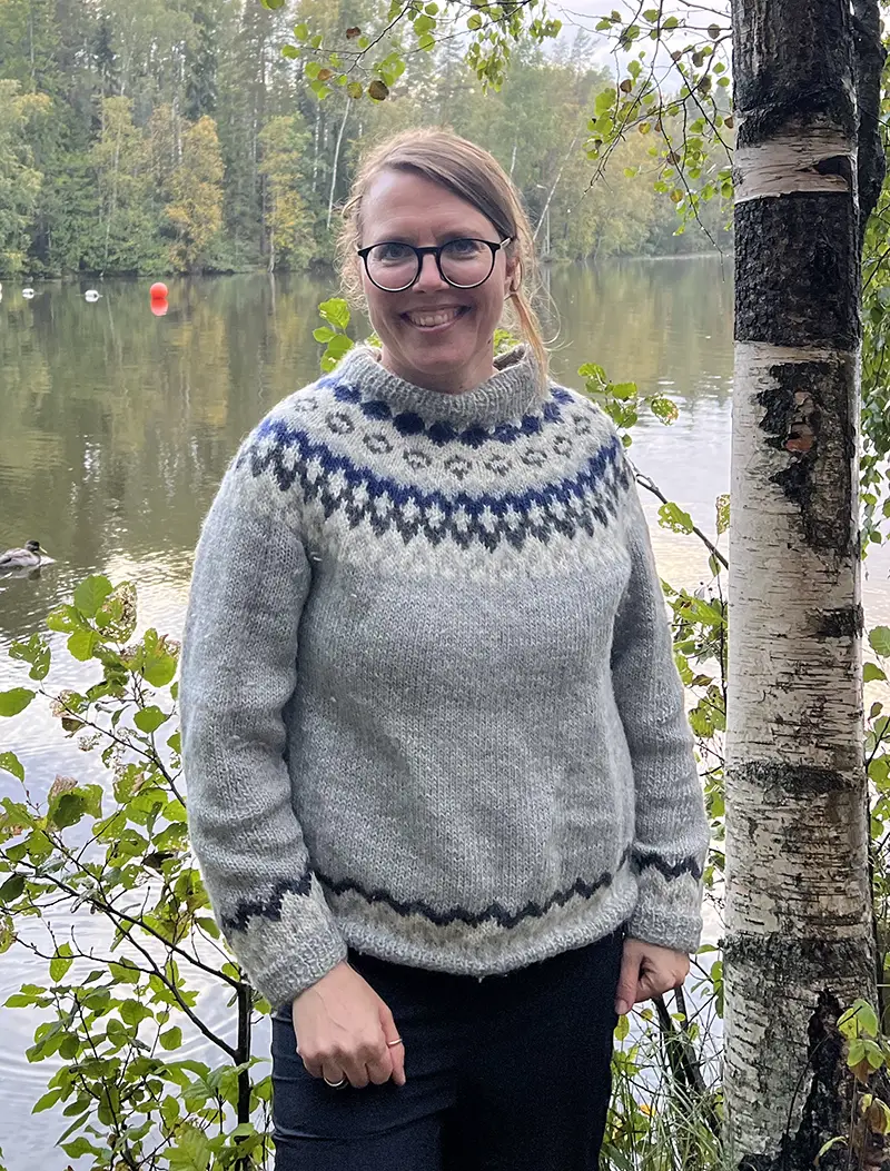 Eeva Eronen-Rasimus A woman in a sweater smiles at the camera next to a birch tree, with a lake in the background.