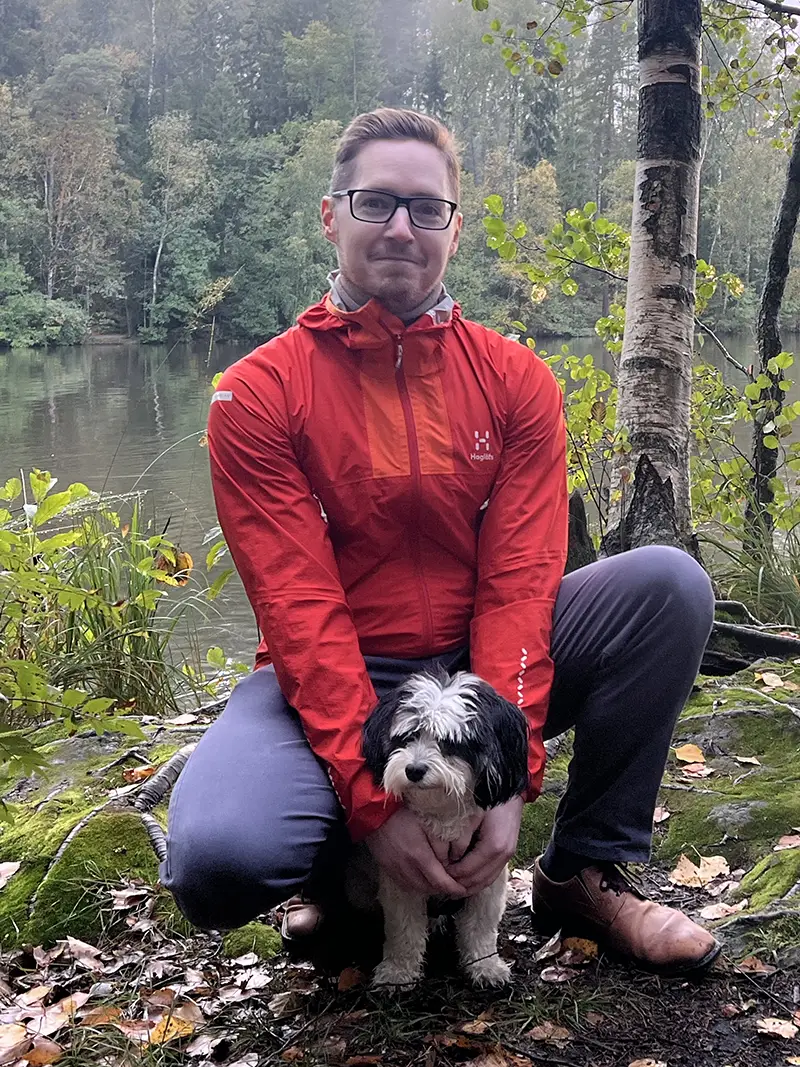 Johannes Cairns A man in a orange-red jacket looks at the camera with a small dog, with a lake in the background.