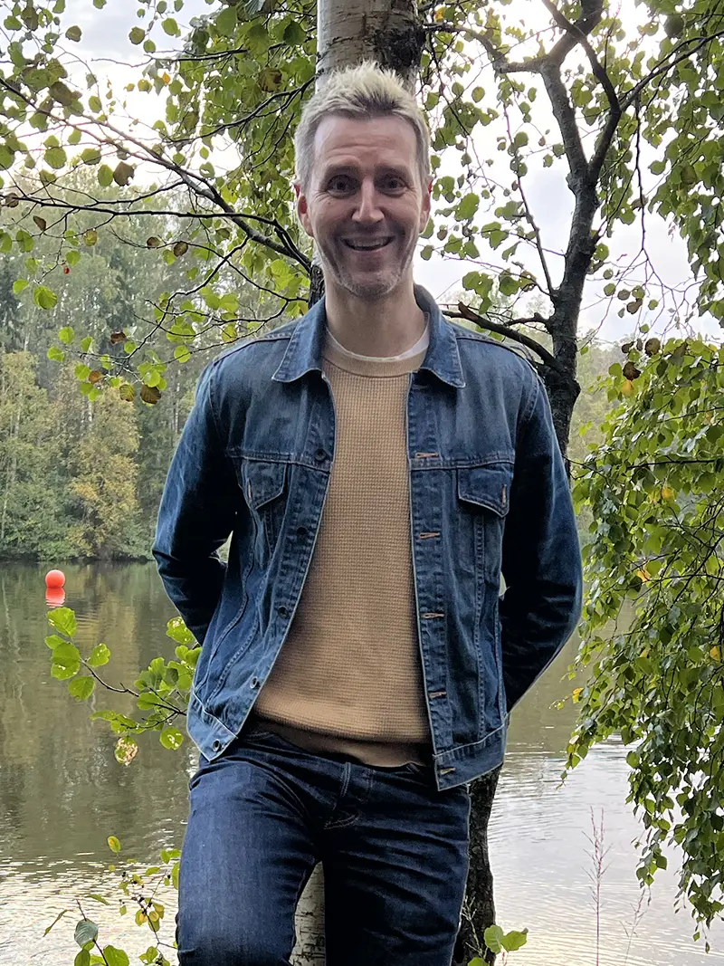 Niko Urho A man in a denim jacket smiles at the camera, leaning against a birch tree with a lake in the background.