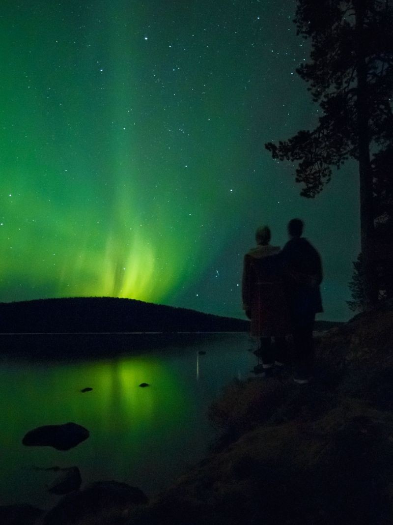 wo dark figures watching the Northern Lights, with a calm lake in front of them and a fell rising behind it.