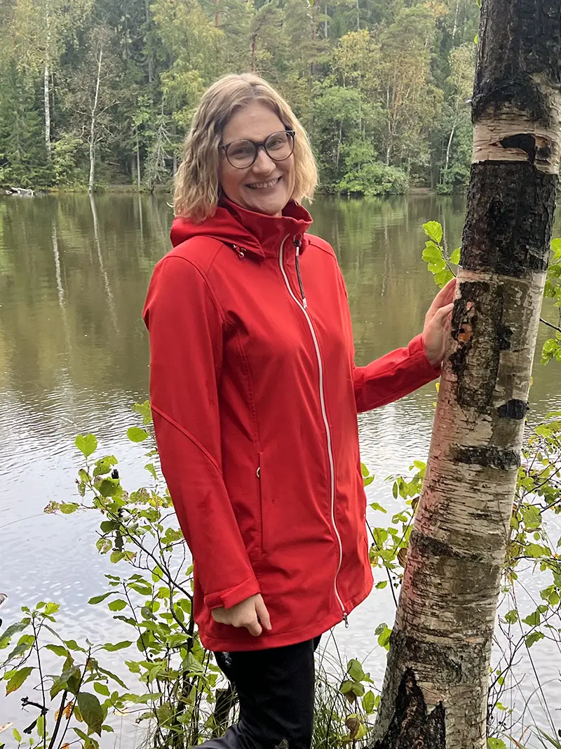 Päivi Suihkonen A woman in a red coat poses next to a birch tree with a lake in the background.