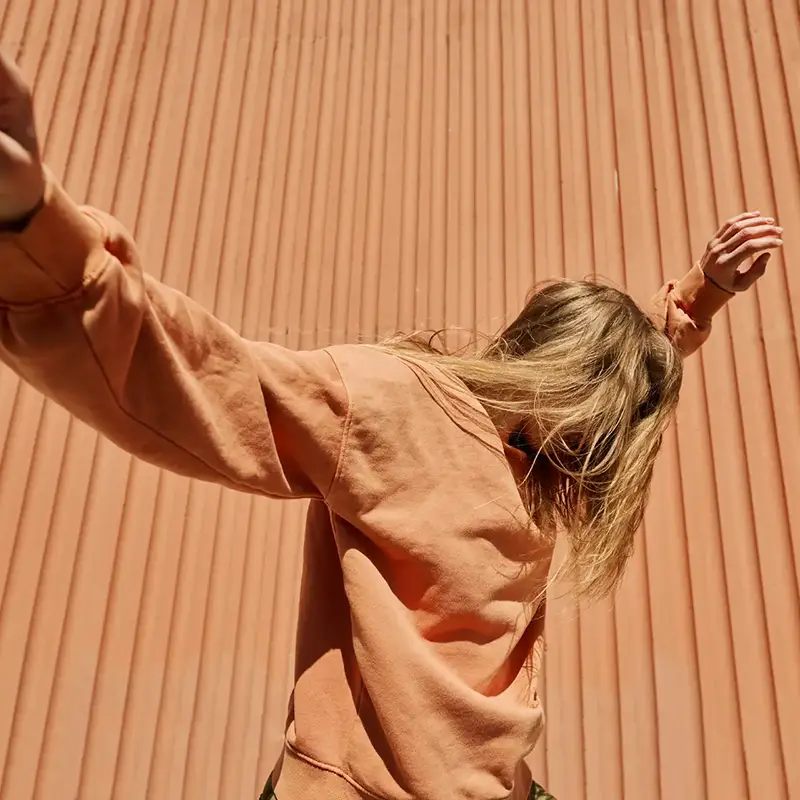 A woman in an orange shirt in front of an orange wall raises her hands in the air, with her hair covering her face.